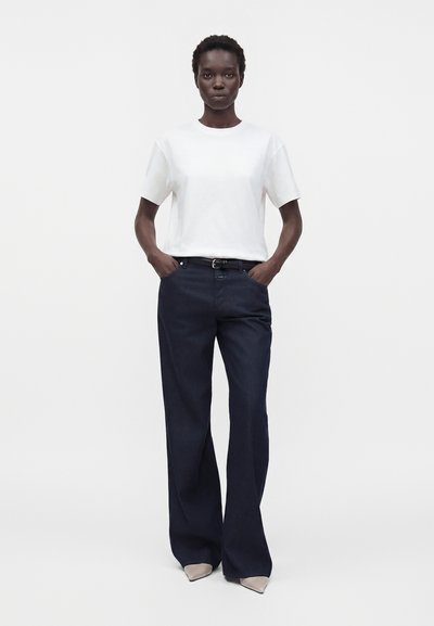 Woman with short hair stands with hands in pockets, wearing a white short-sleeve top, dark wide-leg pants, black belt, and beige pointed shoes.