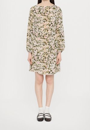 Mujer con un vestido floral de manga larga hasta la rodilla con cinturón, calcetines blancos y zapatos Mary Jane negros, de pie contra un fondo blanco.