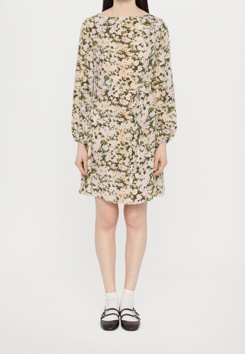 Femme portant une robe fleurie à manches longues, longueur genou, avec une ceinture, des chaussettes blanches et des chaussures Mary Jane noires, debout devant un fond blanc.
