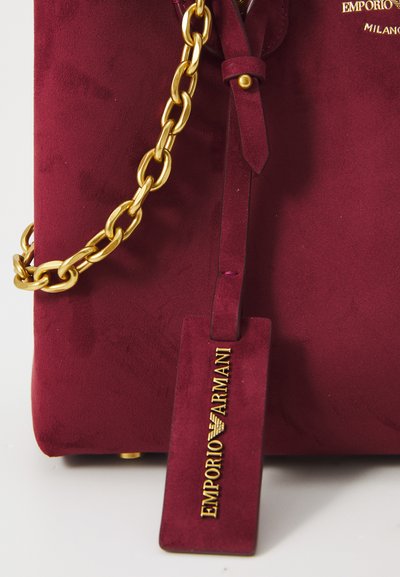 Burgundská zamšová taška so zlatým reťazovým popruhom a dekoratívnou visačkou s nápisom "Emporio Armani" v tučnom písmene. Hladká textura a minimalistický dizajn.