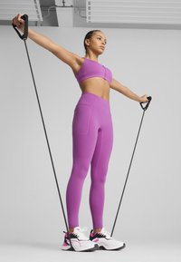 Sutien sport violet și colanți cu talie înaltă, material texturat, design ajustat. Modelul folosește o bandă de rezistență neagră. Pantofi sport albi și roz.