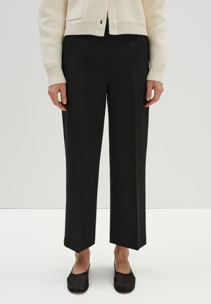 Persoon die zwarte, op maat gemaakte cropped broek draagt, een crèmekleurig vest met knopen en zwarte platte schoenen, staand op een witte vloer.