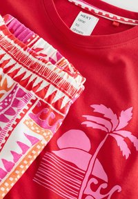 Červený odev s potlačou ružového palmového stromu a slnka vedľa bielej, ružovej a oranžovej vzorovanej tkaniny s elastickým pásom, zobrazuje odevnú štítok.