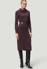 Braunes Strickkleid mit schulterfreier Ausschnittlinie, langen Ärmeln und tailliertem Bund. Kombiniert mit kniehohen, glänzenden schwarzen Stiefeln.