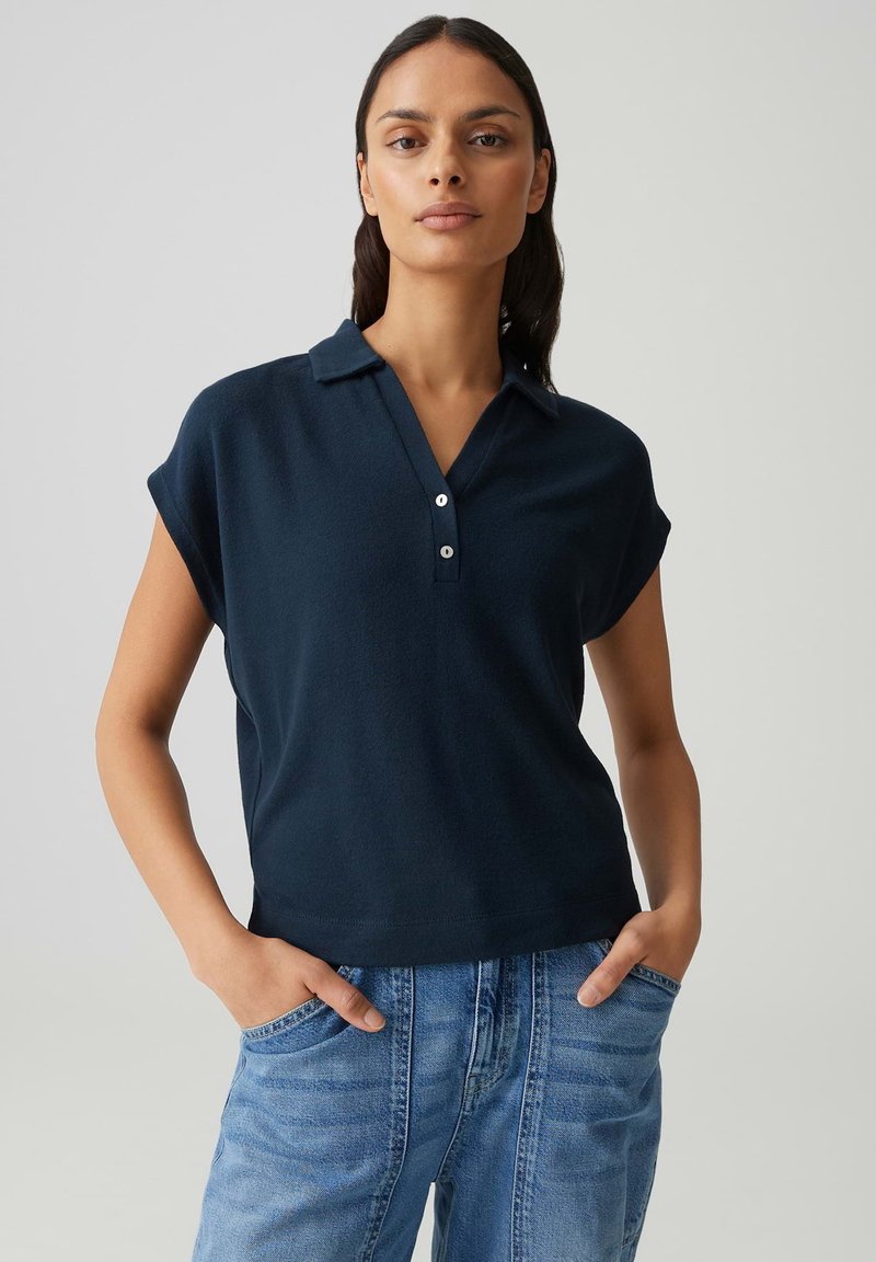 Femme debout, les mains dans les poches de son jean bleu, portant une chemise bleu marine à col et manches courtes, sur un fond clair uni.