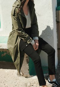 Wodoodporna kurtka w kolorze oliwkowej zieleni, wyposażona w kaptur i przednie kieszenie, zestawiona z czarnymi, wysokim stanem spodniami z dżinsu oraz białymi trampkami.