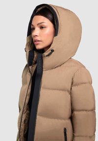 Chaqueta acolchada marrón claro con una gran capucha, cierre de cremallera y acentos negros, que presenta una textura suave y elementos de diseño acolchados.