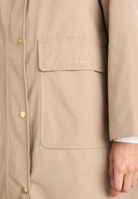 Beige waterdichte jas met een ontspannen pasvorm, voorzien van een grote zak en gouden drukkers. Subtiel logo geborduurd op de zak.