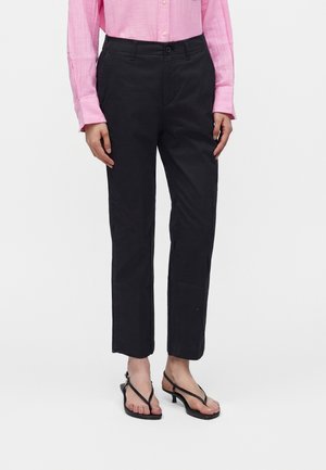 Άτομο που φοράει μαύρο ίσιο παντελόνι, ροζ μακρυμάνικο πουκάμισο με κουμπιά και μαύρα σανδάλια με τακούνι, στέκεται μπροστά σε λευκό φόντο.