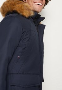 Muž se usmívá, má na sobě tmavě modrou zimní bundu s kapucí z kožíšku a zipem vpředu, na jednoduchém bílém pozadí.