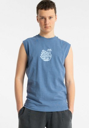 Młody mężczyzna w niebieskiej koszulce bez rękawów z napisem „Stay Sunny” i grafiką palmy, ręce w kieszeniach ciemnych spodni, neutralne tło.