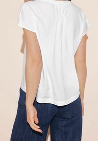 Maglietta bianca a maniche corte con una vestibilità comoda, caratterizzata da un dettaglio plissettato sul retro. Abbinata a pantaloni di denim blu scuro.