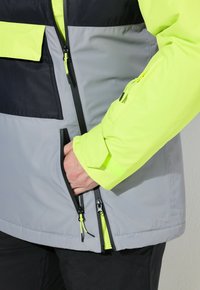 Osoba ubrana w kurtkę odblaskową z czarnymi, szarymi i neonowo żółtymi panelami, z ręką włożoną do zapinanej na zamek kieszeni bocznej.
