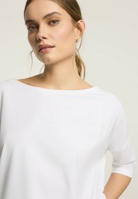 Biała, luźno dopasowana bluzka z szerokim dekoltem i rękawami trzyczwartowymi, wykonana z miękkiego materiału o gładkiej fakturze.