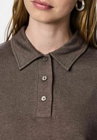 Izbliza prikaz osobe koja nosi smeđu pamučnu polo majicu sa tri dugmeta i srebrnim okruglim naušnicama, prikazujući vrat i donji deo lica.