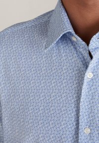 Koszula w niebieskie wzory z drobnymi kwiatowymi motywami w różnych odcieniach niebieskiego, z kołnierzykiem i zapięciem na guziki. Zauważalna tekstura bawełnianego materiału.