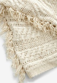 Cremefarvet strikplaid med et struktureret vævet mønster, der har frynset kanter og et blødt, chunky design.