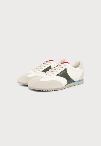 Paire de baskets blanches et beiges avec des panneaux latéraux noirs, une doublure intérieure rouge, une bande bleue sur la semelle, et le logo "Paul Smith" sur le côté extérieur.