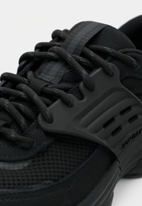 Czarny sportowy but z siateczką i teksturowanym materiałem, z grubymi sznurowadłami, formowanym oczkiem i subtelnym logo po boku.