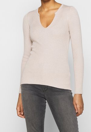 Woman wearing a beige ribbed long-sleeve V-neck sweater paired with dark gray jeans, standing against a plain background.