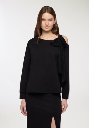 Donna che indossa un top nero a maniche lunghe con una spalla scoperta e un grande fiocco di pizzo, abbinato a una gonna nera con uno spacco frontale.