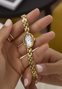 Orologio Timex dorato con quadrante ovale bianco, numeri romani neri e bracciale in metallo dorato. Mostrato in una mano.