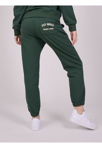 Groene joggingbroek van zacht materiaal, met een zak en beige tekst "PXP SQUAD PROJECT X PARIS" en elastische boorden. Draagt een paar witte sneakers.