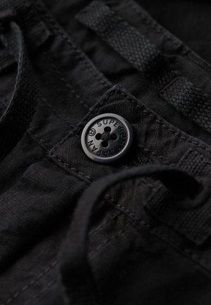 Sort stof med en struktureret finish, prydet med en sort knap prægnet med "Superdry" og en snøre detalje.