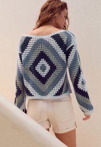 Sweter na szydełku z długimi rękawami, z wzorem diamentowym w odcieniach niebieskiego, zielonego i białego. Przecięty w talii, zestawiony z białymi szortami.