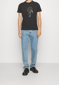 Camiseta de algodón negra con un diseño gráfico brillante de un personaje. Combinada con unos jeans de corte relajado en azul claro y zapatos negros.