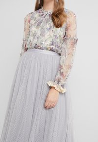 Blouse fleurie avec manches transparentes et poignets à volants, associée à une jupe en tulle gris clair ornée d'un motif à pois texturé.