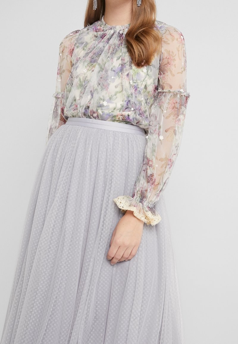 Blouse fleurie avec manches transparentes et poignets à volants, associée à une jupe en tulle gris clair ornée d'un motif à pois texturé.