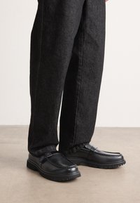 Sorte lærskofe med struktureret overflade, med hævede søm-design og en solid gummisål, båret sammen med mørke denimbukser.