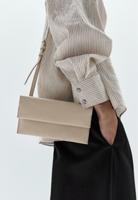 Borsa structured beige con due pannelli, texture liscia e tracolla sottile, abbinata a una camicia a righe crema e una gonna di tessuto nera.