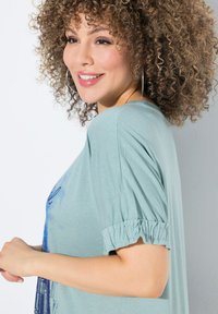Top de jersey verde claro con un corte holgado, mangas cortas fruncidas y un diseño gráfico azul en la parte frontal. Textura suave y estilo casual.