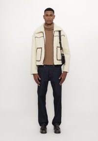 Veste en shearling couleur crème avec des accents bruns, pull en tricot torsadé marron, jeans en denim foncé et chaussures brunes. Silhouette droite, matériaux texturés.