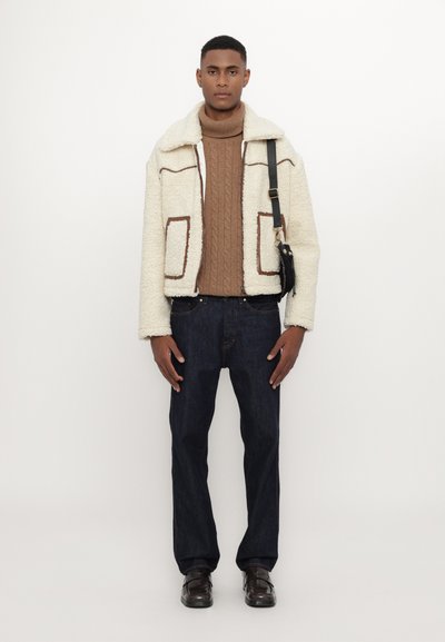 Cream-colored shearling jacket with brown accents, brown cable-knit sweater, dark denim jeans, and brown shoes. Straight silhouette, textured materials.