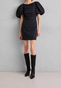 Svart figursydd klänning med puffärmar, texturerad tyg och rynkade detaljer. Kombinerad med knähöga svarta klackstövlar. En enkel betongbakgrund.