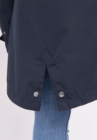 Marinblå regnjacka med en strukturerad yta, med en spatial ryggslits och silverfärgade tryckknappar i nederkant.