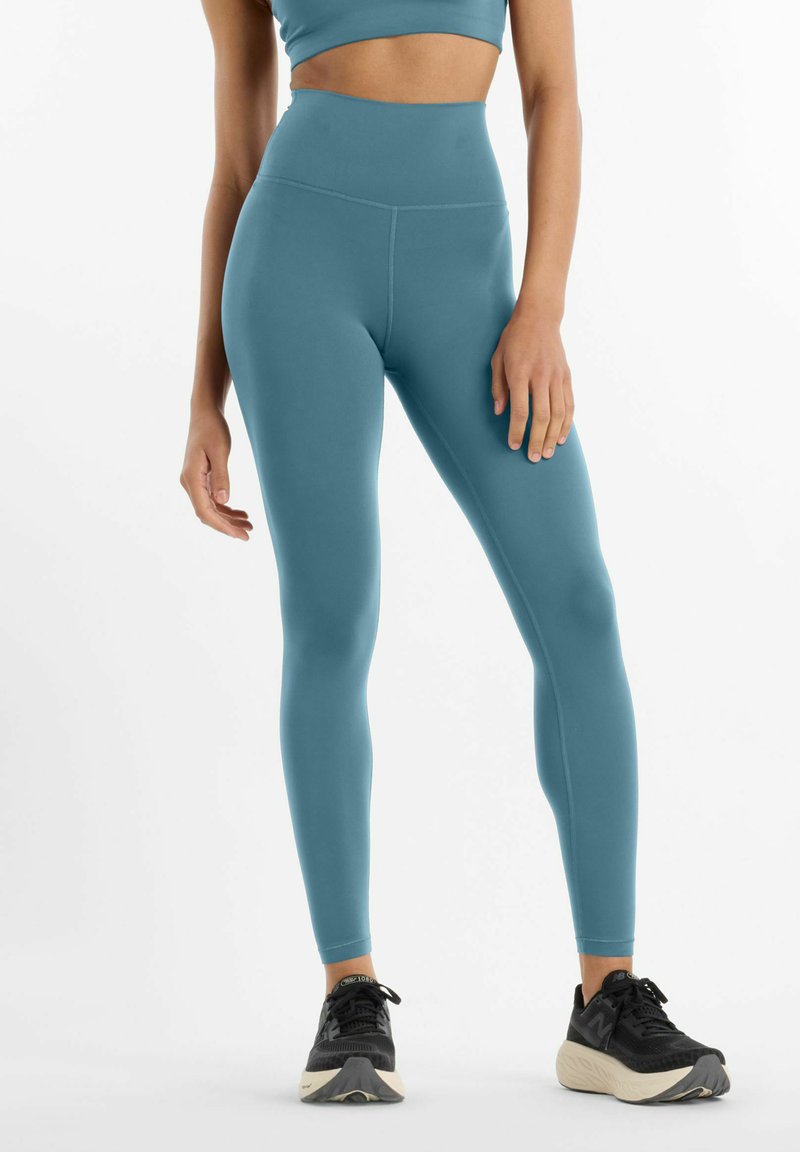 Femme portant des leggings taille haute couleur sarcelle et un crop top assorti, debout avec des chaussures de course noires sur un fond blanc.