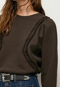 Sudadera marrón con acentos texturizados y mangas abullonadas, con un cuello de canalé y un corte relajado, combinada con un cinturón oscuro y jeans de mezclilla.