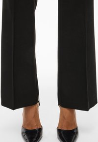 Underkropp med svarte vide bukser og spisse svarte lær-sko mot en hvit bakgrunn.