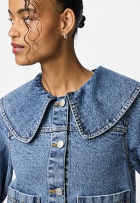 Gaiši zils džinsūķis ar lielu noapaļotu apkakli, priekšējo pogu aizdari un divām apakšējām kabatām ar redzamu šuvi.