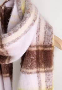 Brun och krämvit rutig halsduk, gjord av mjuk ull. Har en mjuk struktur med en ljusgul accent i mönstret. Omsvept på en galge.
