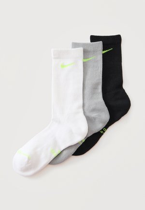 Trois chaussettes Nike crew blanches, grises et noires avec un logo et texte vert fluo, disposées en chevauchement sur un fond clair.