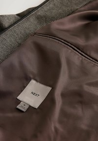 Tela gris texturada con un forro marrón suave. Presenta una etiqueta con la inscripción "NEXT," tamaño "M," y una etiqueta que indica "Hecho en Portugal."