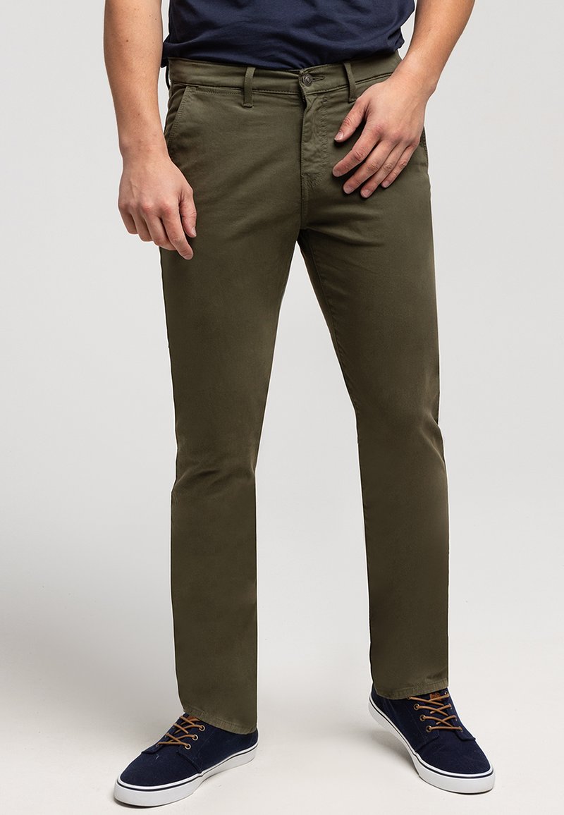 Oliwkowe chinosy o wąskim kroju, z zapięciem na guzik i przednimi kieszeniami, w połączeniu z granatowymi butami i ciemnoniebieską koszulą.