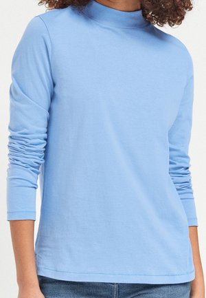 Personne portant un haut bleu clair à col montant et manches longues froncées, avec un jean bleu, sur un fond uni.