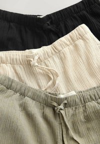 Tre par texturerade byxor med dragsko i svart, beige och oliv, staplade med synliga elastiska midjeband och tygetiketter.