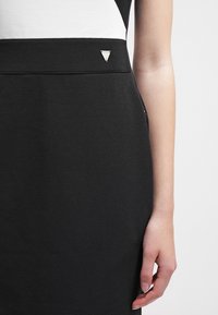 Jupe noire texturée avec une petite épingle en argent en forme de triangle sur la taille, portée avec un haut blanc, montrant le bras droit et la main détendue.